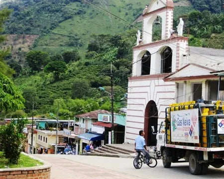 A small Colombian town with a white church featuring a bell tower, surrounded by lush green mountains. A man rides a bicycle past a parked truck on the main street, with shops and houses lining the hillside.