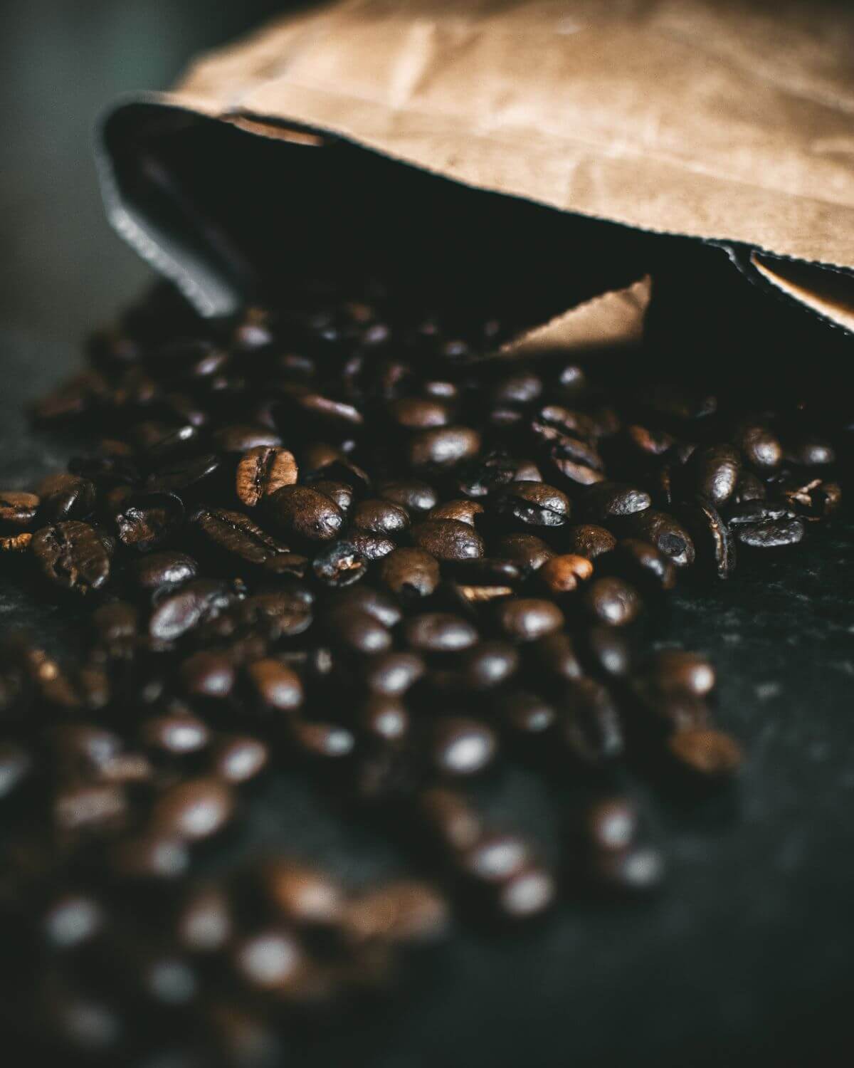Roasted coffee beans spilling out of a brown paper bag on a dark surface