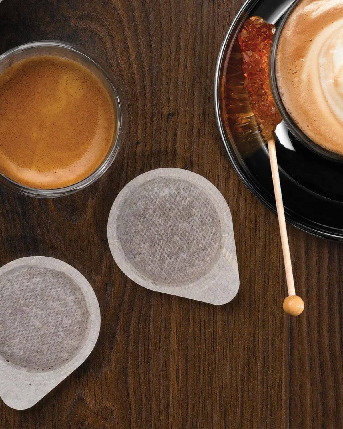 Two ESE coffee pods on a wooden surface with a cup of coffee in the background.