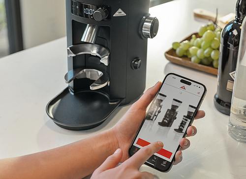 Person adjusting Mahlkönig coffee grinder settings using the mobile app on a smartphone in a modern kitchen.