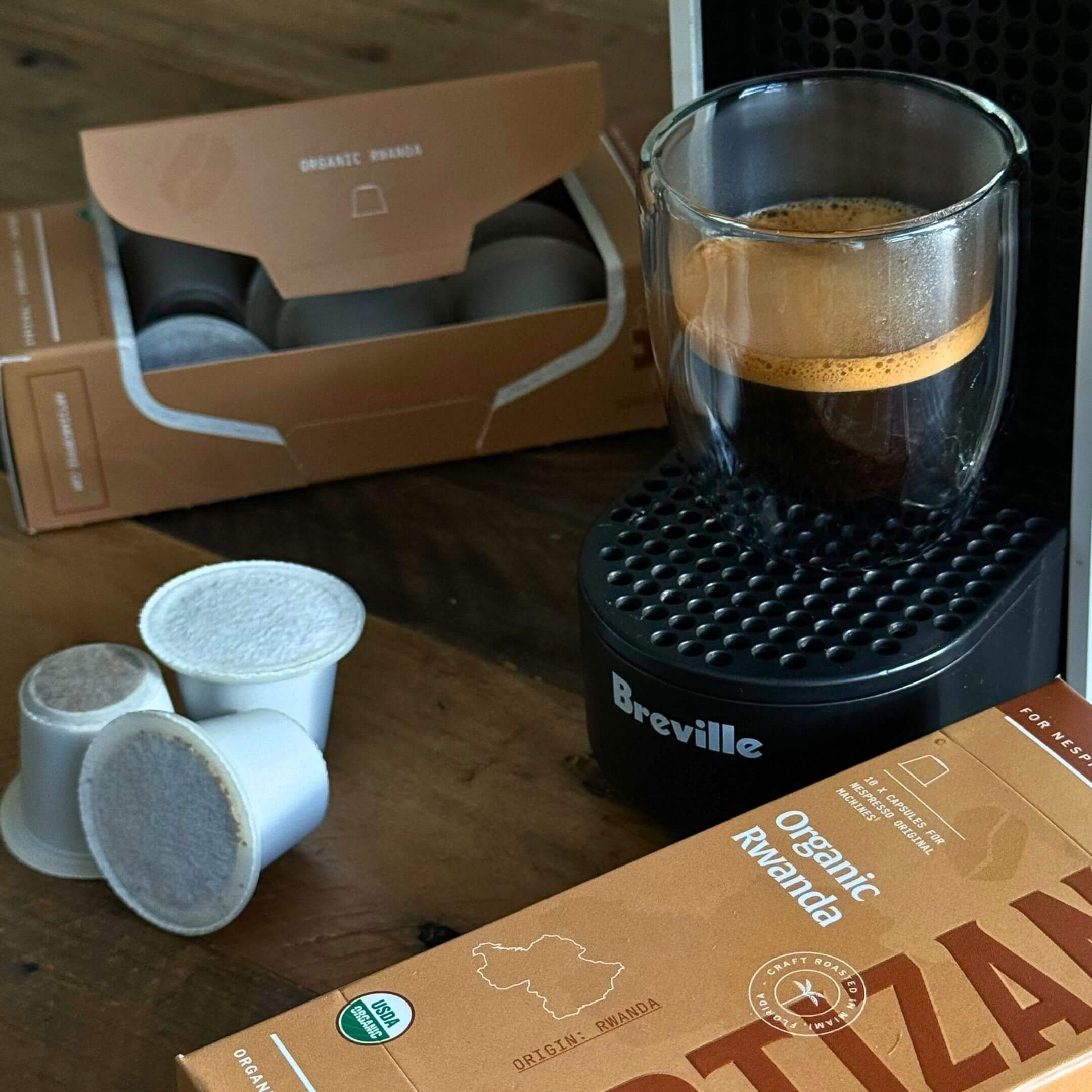 Image of Coffee on a Breville Coffee Machine with Packaging of Organic Rwanda and Capsules for Nespresso Original Machine on Top of a Wooden Table.