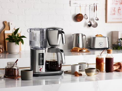 Lifestyle kitchen scene with The Luxe Brewer Glass automatic drip coffee maker on countertop surrounded by mugs, pastries, and coffee accessories, highlighting Breville glass carafe design.