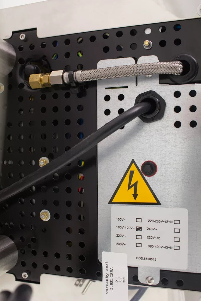 Close-up of Bezzera Mitica V2 HX espresso machine back panel with cables, metal conduit, warning sign, and voltage label.
