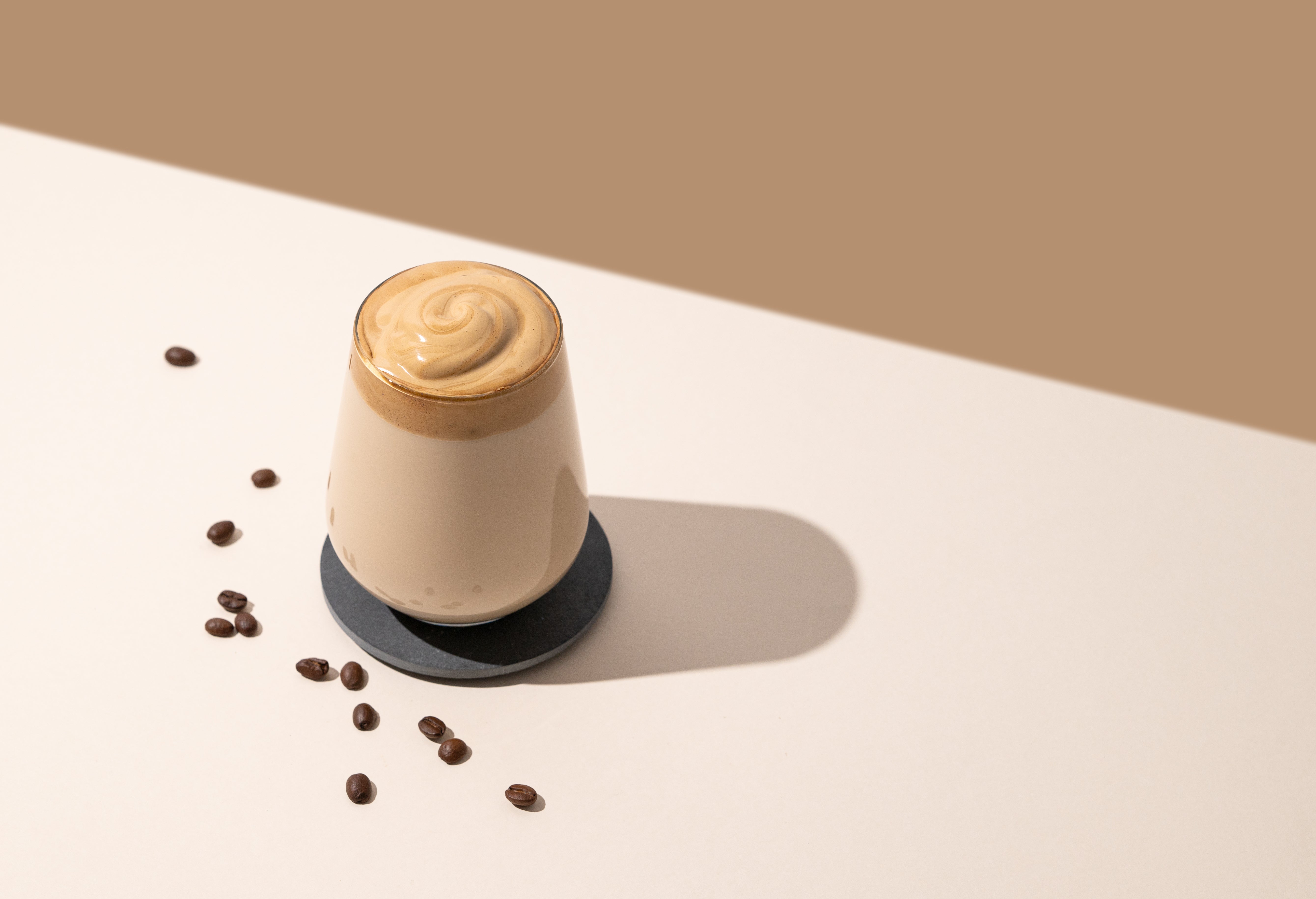 Dalgona coffee whipped instant vegan latte with frothy swirl served in glass on coaster with roasted coffee beans minimalist background