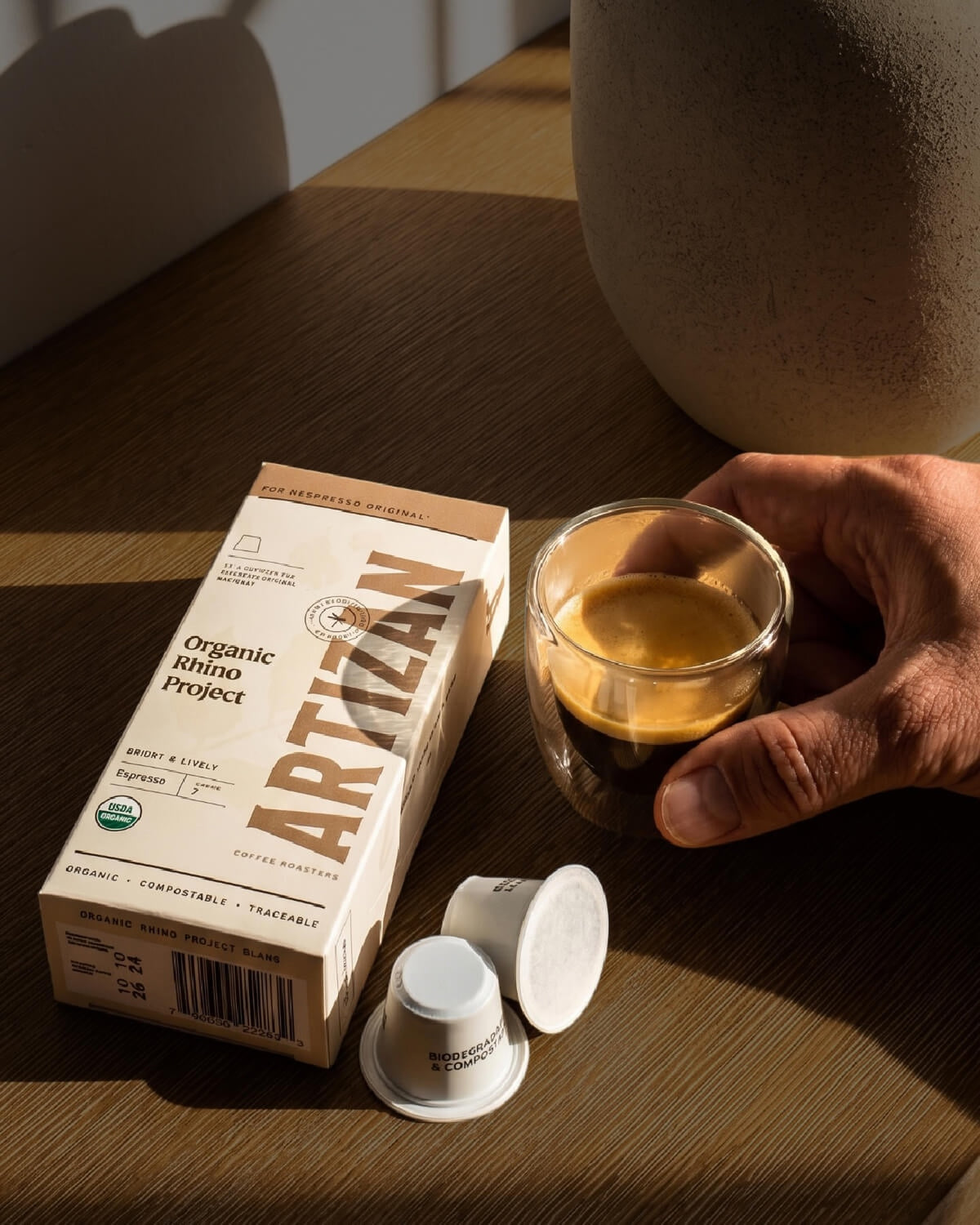 A hand holds a small glass of espresso beside a box of Artizan Organic Rhino Project espresso capsules and two capsule lids on a wooden table.