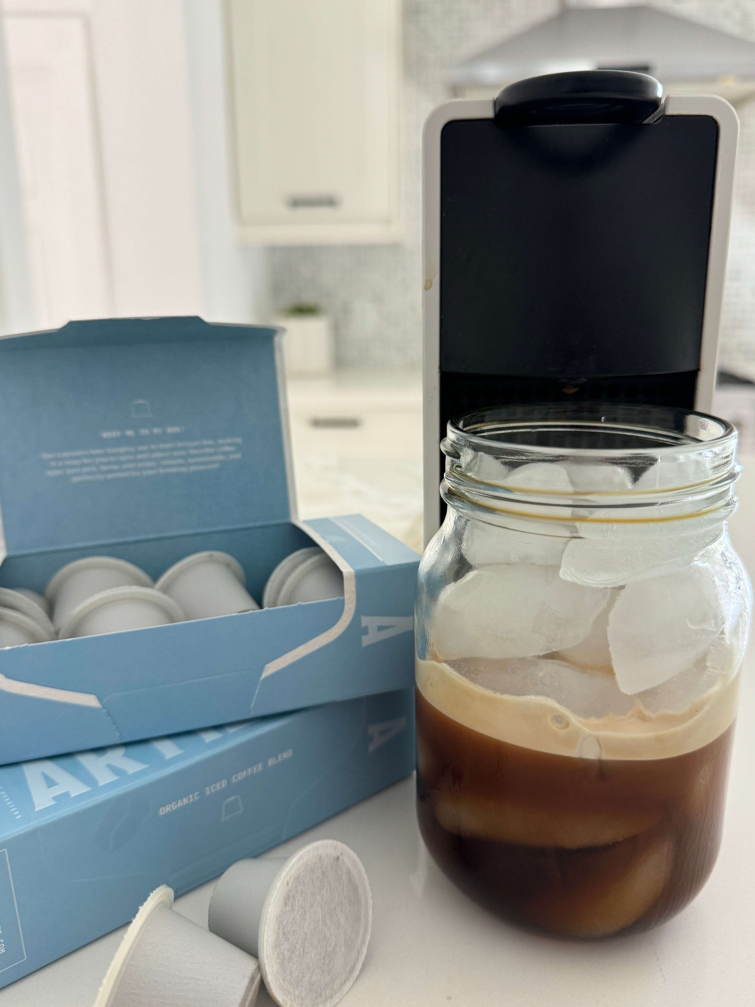 Freshly brewed organic iced coffee in a glass jar with ice, beside Artizan iced coffee pods and a pod coffee machine on a modern kitchen counter