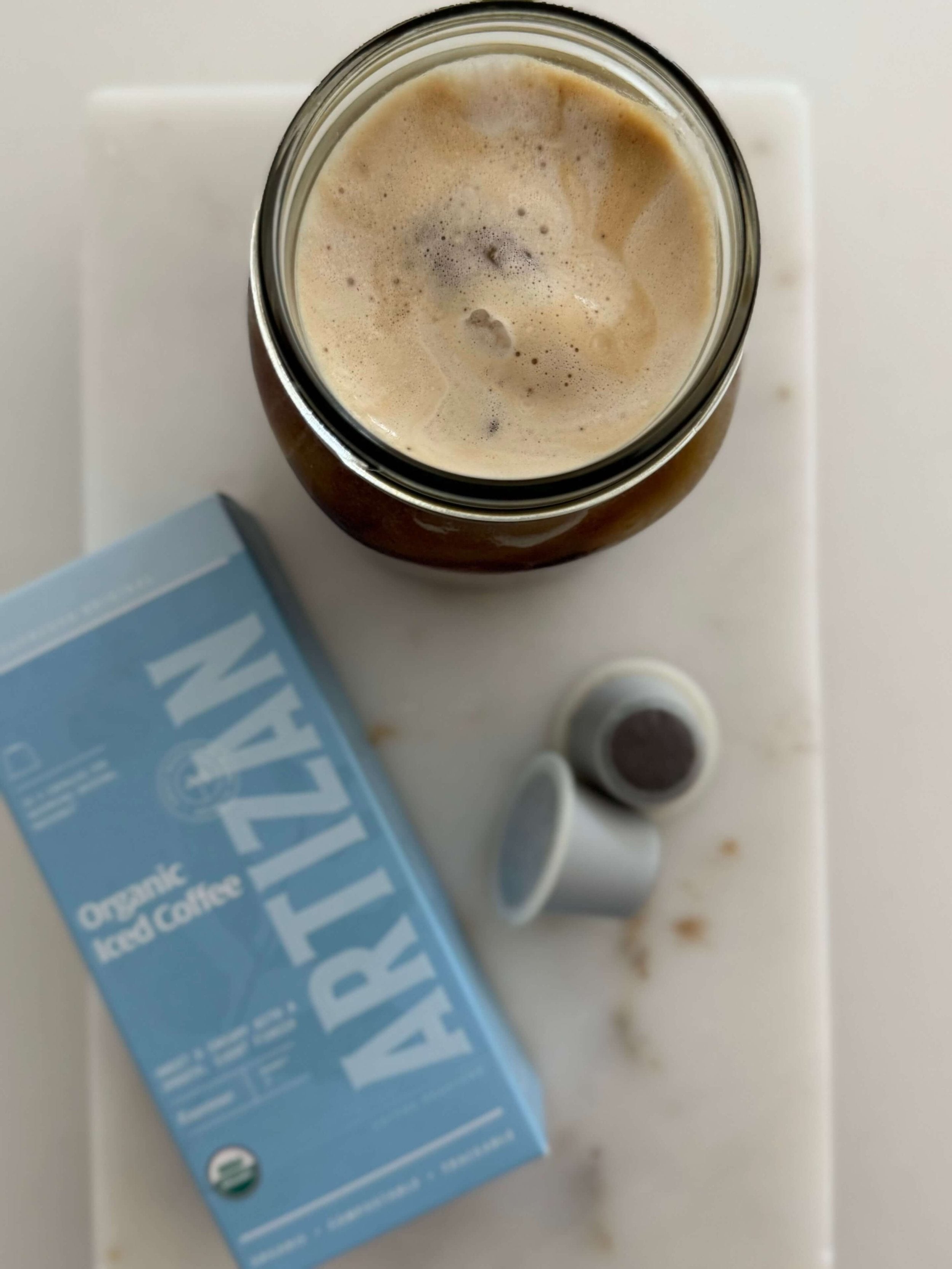 Glass of organic iced coffee with creamy foam on top, placed beside Artizan Organic Iced Coffee pods and packaging on a marble surface.