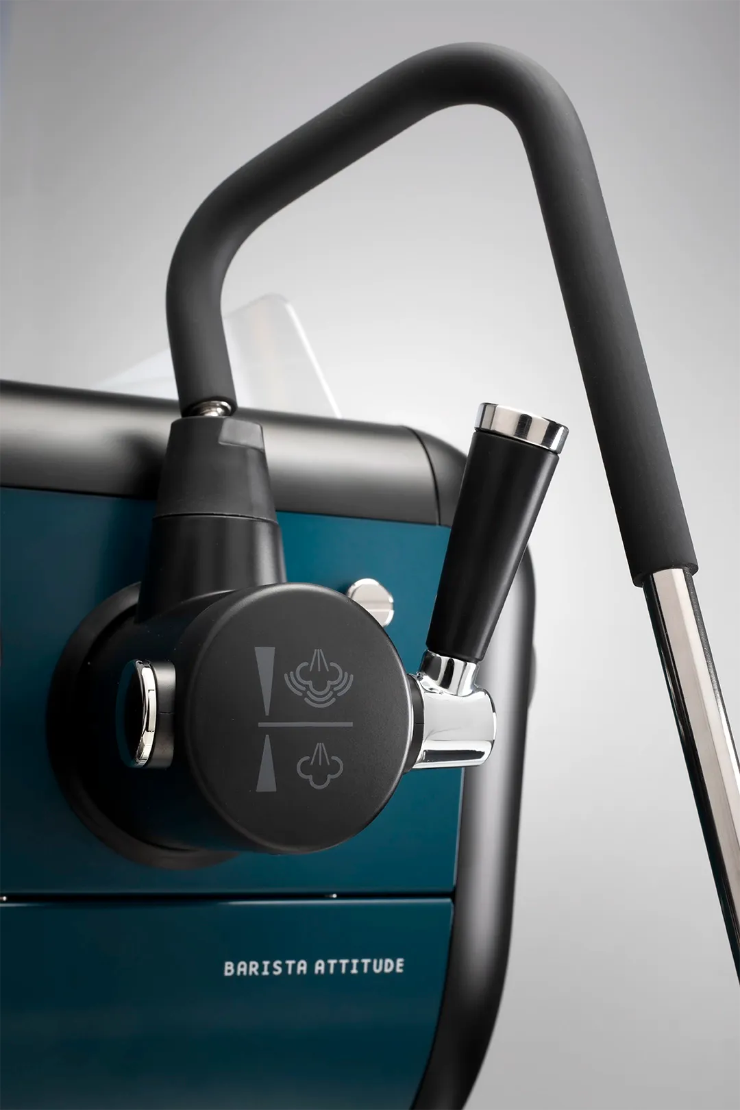 A close-up view of a modern espresso machine’s steam wand and control knob, featuring a matte black handle, chrome accents, and labeled steam icons. The machine’s body is teal, with the words “Barista Attitude” visible on the front.