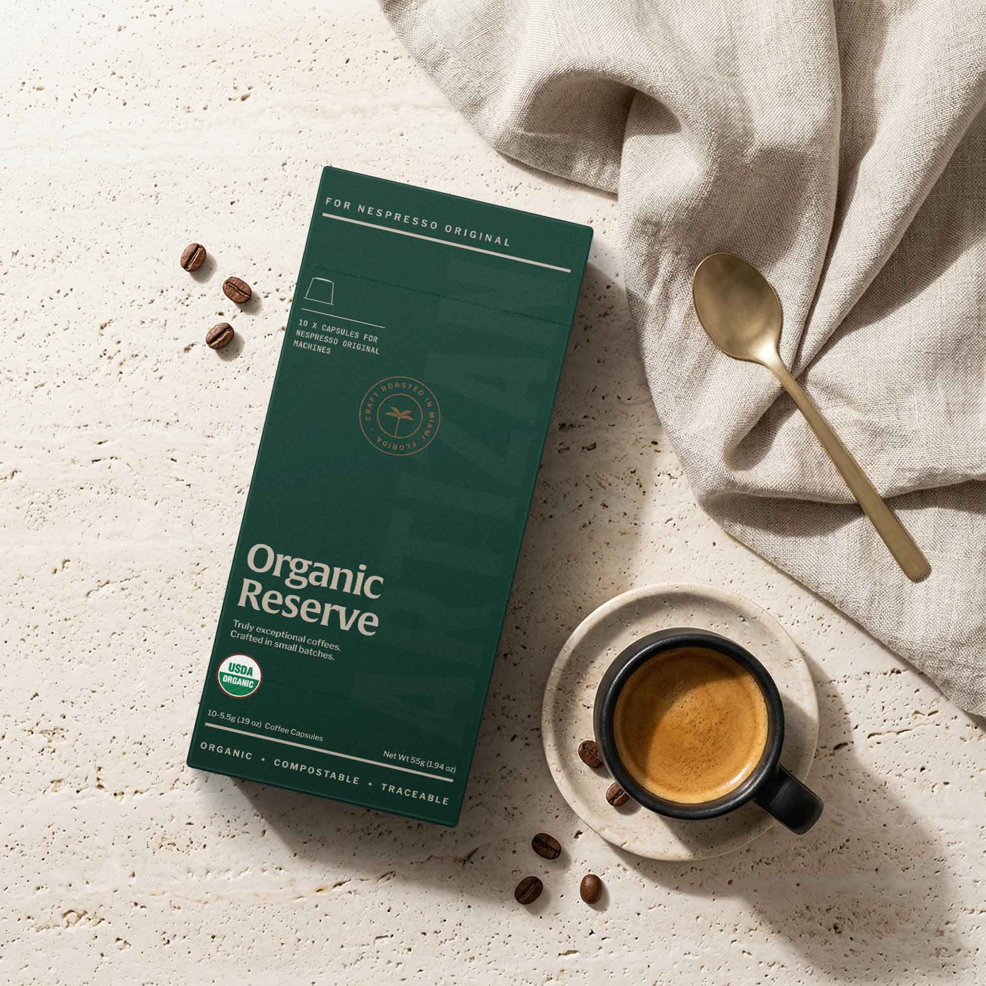Organic Reserve coffee capsules for Nespresso Original displayed with freshly brewed espresso, scattered coffee beans, and gold spoon on a textured travertine background