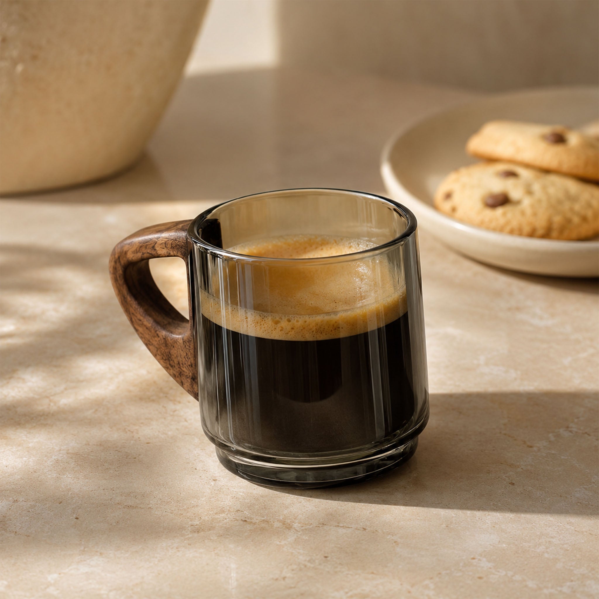 Smoky gray glass espresso cup with a wooden handle filled with espresso on a sunlit countertop beside cookies.
