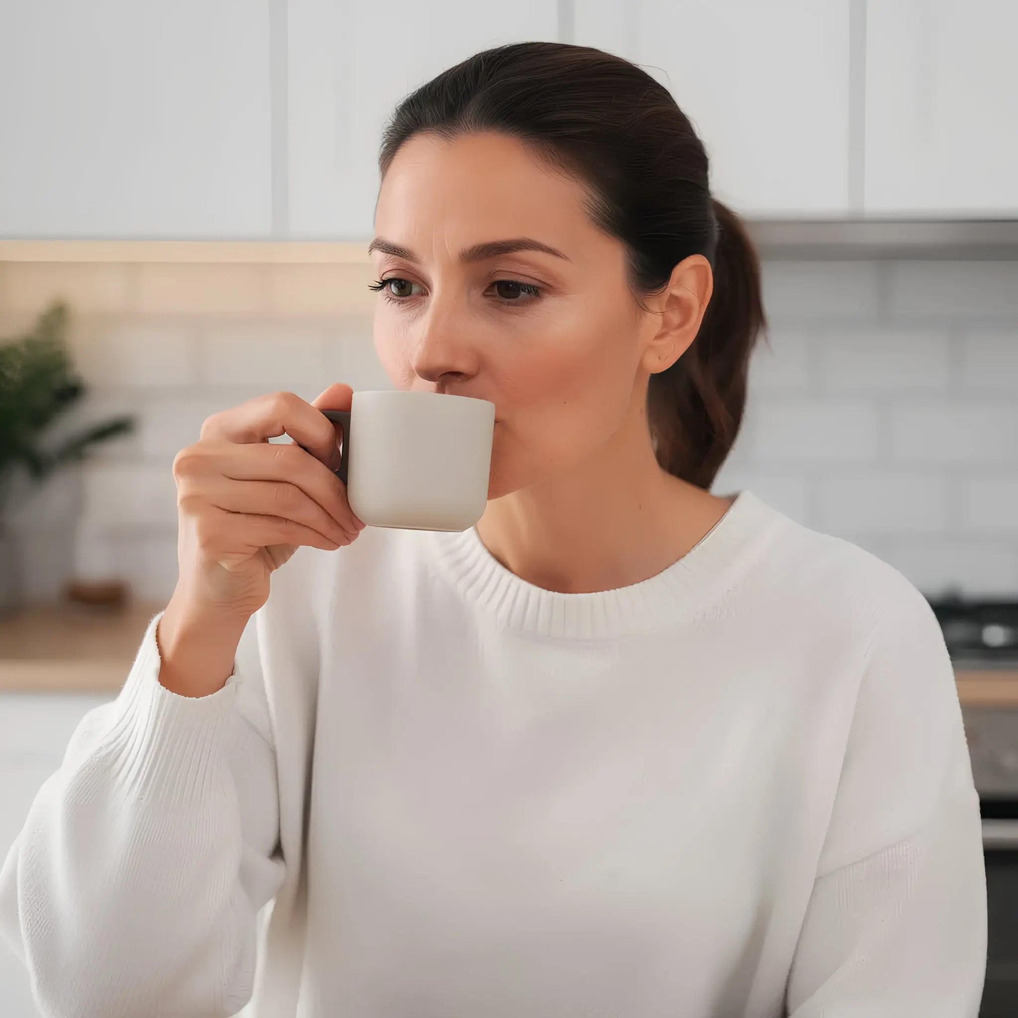 Woman in white sweater sipping from a stylish gray coffee cup in a modern kitchen setting.